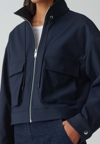 Marinblå kort jacka med hög krage, silverfärgad dragkedja, två framfickor och justerbara dragsko. Tillverkad av slät, texturerad tyg.