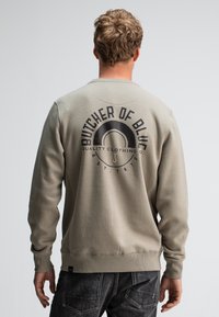 Grijze sweatshirt met lange mouwen, geribbelde manchetten en zoom. Heeft een zwart grafisch logo op de achterkant, dat de merknaam en het oprichtingsjaar benadrukt.
