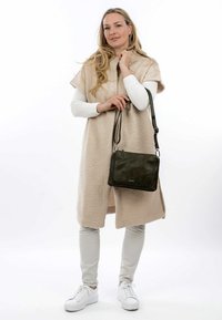 Poncho in pile beige sopra una maglietta a maniche lunghe bianca, abbinato a pantaloni grigi chiari e sneaker bianche. È tenuta una borsa a tracolla verde oliva.