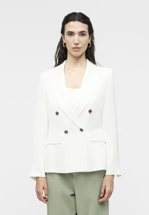 Ženska, oblečena v bel dvorezni blazer z zlatimi gumbi in svetlo zelenimi hlačami, stoji na preprostem svetlem ozadju.