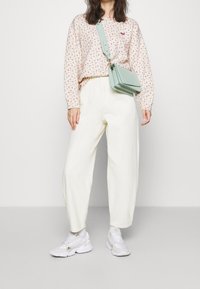 Donna che indossa un sweatshirt bianco con stampa floreale, pantaloni bianchi larghi, sneakers bianche e una borsa a tracolla verde menta contro uno sfondo neutro.