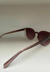 Ronde zonnebril met een doorschijnend donker bordeauxrood montuur en donkere glazen, voorzien van een slanke, gebogen pootjesontwerp. Geëtiketteerd met merknaam en UV-beschermingscategorie.