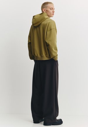 Olívazöld, kapucnis, túlságosan bő pulóver bordázott mandzsettákkal, laza fekete nadrággal és masszív fekete lábbelivel párosítva.