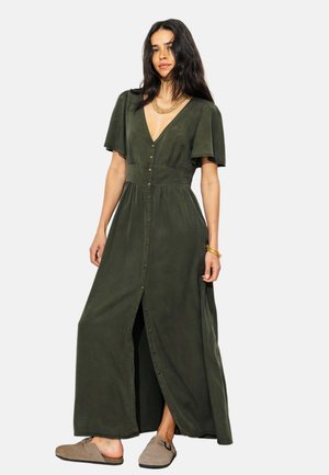 Femme debout, portant une longue robe vert olive à boutons sur le devant avec des manches courtes, associée à des chaussures beiges sans lacets et des bijoux en or.