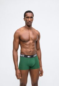 Μυώδης άνδρας με κοντά σγουρά μαλλιά, βρίσκεται μπροστά, φοράει πράσινα μπόξερ Lacoste με μαύρη μέση σε απλό λευκό φόντο.