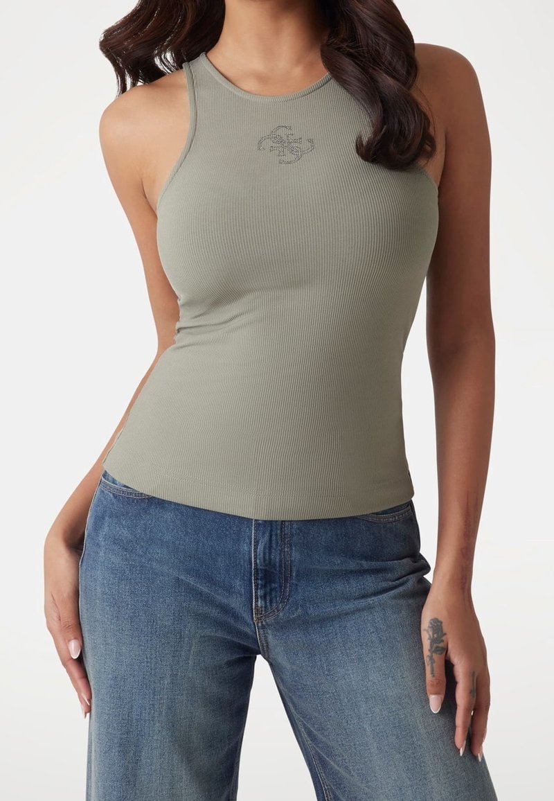 Femme portant un haut côtelé sans manches vert clair et un jean bleu, avec un tatouage sur le doigt de la main gauche et des cheveux foncés lâchés.