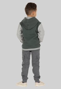 Groene hoodie met grijze mouwen, gecombineerd met grijze cargojoggers. Kenmerkend zijn de gestreepte tailleband en zakken op de broek. Basis lichte schoenen.