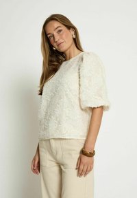 Lace blouse in ivory, featuring puff sleeves and a textured floral design. Paired with light beige trousers and a bangle on the wrist.
