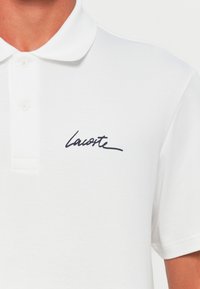 Polo blanc en coton, doté d'un col à trois boutons et d'un logo "Lacoste" brodé en bleu marine sur la poitrine gauche.