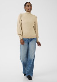 Nais, kes seisab beežis pikkade varrukatega kõrge kaelusega kampsunis, avarates sinistes teksastes ja mustades kingades, ühtlase valge tausta ees.