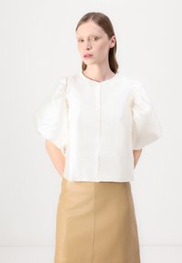 White satin blouse with puffed sleeves, round neckline, and central front closure. Worn with a tan leather skirt, smooth texture.