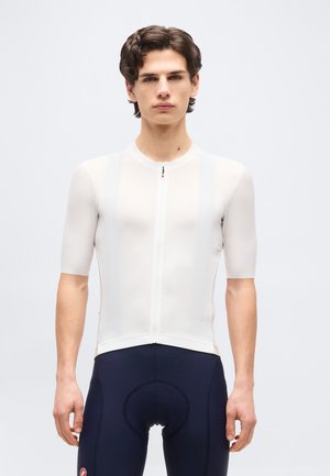 Νεαρός άνδρας που φοράει εφαρμοστό λευκό ποδηλατικό μπλουζάκι με κοντά μανίκια και μπλε σκουρόχρωμο ποδηλατικό σορτς με επένδυση, στέκεται μπροστά σε ένα απλό φωτεινό φόντο.