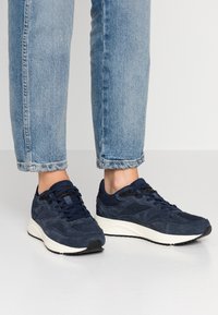 Tmavě modré semišové tenisky s texturovanými panely, černými akcenty a bílými gumovými podrážkami. Vybavené černými tkaničkami a polstrovaným jazykem.