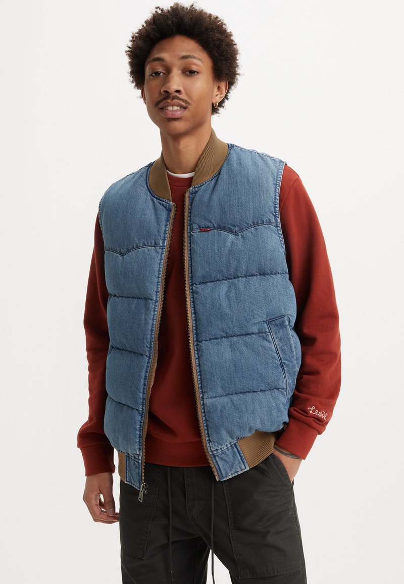Gilet en denim léger bleu avec col côtelé marron et fermeture éclair, présentant des panneaux cousus et deux poches latérales. Superposé à un sweat-shirt rouge.