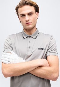 Νέος άνδρας με σταυρωμένα χέρια, φορώντας γκρι πόλο μπλουζάκι BOSS και λευκό γάντι γκολφ στο αριστερό χέρι, μπροστά σε απλό φόντο.
