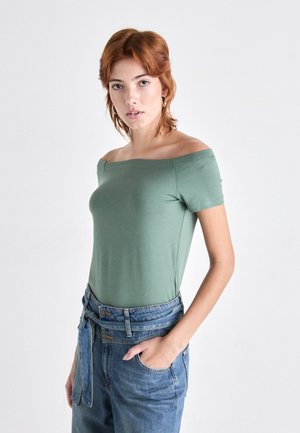 Grønn av-shoulder t-skjorte i lett stoff med korte ermer, parret med høytlivte blå denimjeans med en knyttet belte.