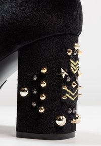 Zapato de tacón alto de terciopelo negro con un tacón cuadrado texturizado. Decorado con tachuelas doradas y negras, y patrones de chevrón bordados en dorado.