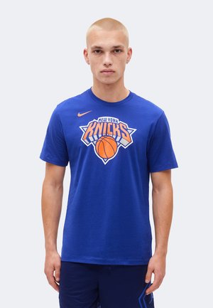 Jeune homme aux cheveux blonds courts portant un t-shirt bleu des New York Knicks et un short bleu assorti, debout devant un fond clair.