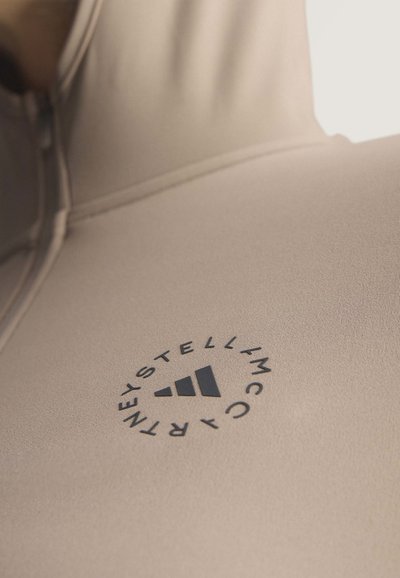 Close-up of beige hoodie fabric with a black circular Stella McCartney Adidas logo on the chest area.