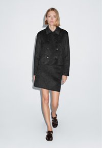 Kvinde med kort blondt hår iført en sort dobbeltradet jakke og en matchende nederdel, der går fremad mod en ensfarvet hvid baggrund.