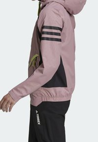 Ljust rosa vindtät jacket med svarta detaljer och tre svarta ränder på vänster arm. Har en dragkedjeficka och elastiska ärmslut.