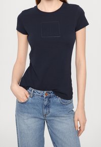 Námořnická modrá tričko s krátkým rukávem vyrobené z bavlny s jemným vyraženým logem na hrudi; spárované se světle modrými džíny.