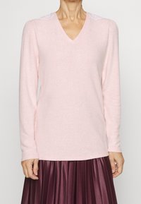 Ljust rosa V-ringad tröja med långa ärmar, med texturerade axelaccenter. Mjuk material, minimalistisk design, kombinerad med en plisserad vinröd kjol.