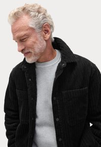 Sort corduroy skjortejakke med en struktureret overflade, der har krave, knaplukning og to frontlommer over en grå ribbestrik sweater.