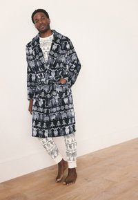 Granatowy szlafrok z białymi wzorami zwierząt i drzew, z miękkiego materiału, z paskiem w talii, noszony na kremowych, wzorzystych spodniach piżamowych, boso na drewnianej podłodze.