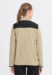 Beige fleecejakke med svart øvre panel og krage. Har lange ermer og svart kant. Myk tekstur, tilpasset passform.