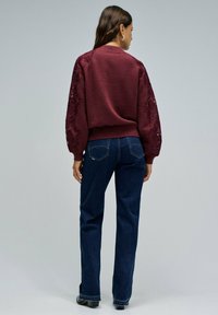 Mujer de pie, vistiendo un suéter burdeos con mangas de encaje y jeans azul oscuro, de espaldas contra un fondo gris claro liso.