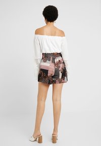 Bílá off-the-shoulder blůza s krajkovými detaily, spojená s černými vzorovanými šortkami s paisley, pruhy a puntíky. Hnědé sandály na podpatku.