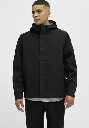 Schwarze wasserdichte Jacke mit Kapuze, Druckknöpfen und zwei Vorderaschen. Hergestellt aus glattem Stoff mit einer lockeren Passform über einem grün gestreiften Hemd.