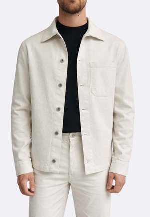 Uomo che indossa una giacca abbottonata color avorio sopra una maglia nera e pantaloni abbinati color avorio, ritratto dal collo in su, in piedi davanti a uno sfondo semplice.