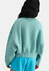 Donna con lunghi capelli scuri che indossa un ampio felpa color teal chiaro e pantaloni corti blu brillante, vista da dietro su uno sfondo bianco.