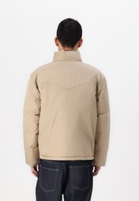 Beige dunsjakke med høy krage, sydd detaljer og en avslappet passform. Båret over mørke denim jeans, som viser en jevn tekstur.