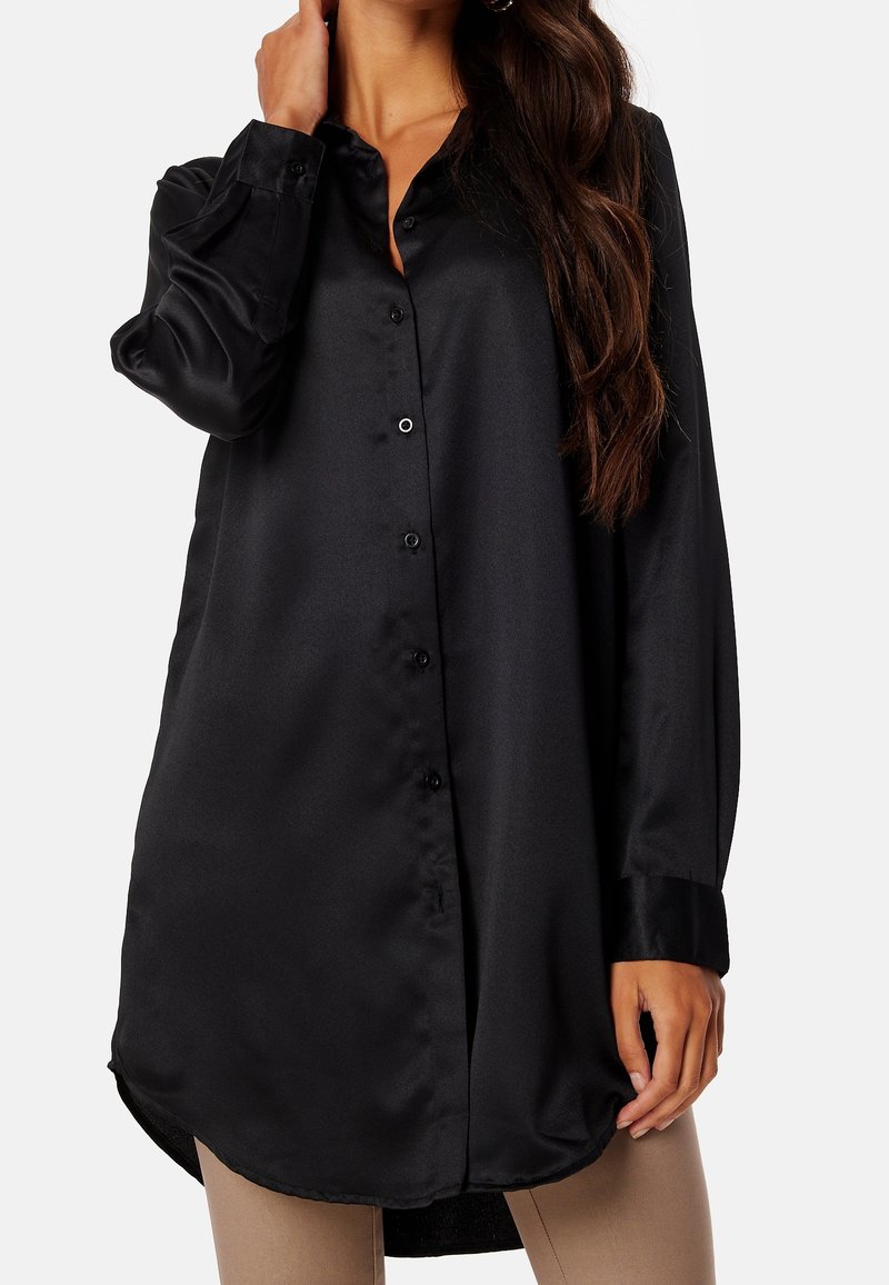 Femme portant une longue chemise en satin noire à boutons avec manches longues et pantalon beige, main près du cou, posture détendue.