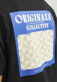 Juodas marškinėliai su mėlynu piešiniu ir užrašu "ORIGINALS COLLECTIVE" virš tekstūrinio smėlio spalvos geometrinio rašto.