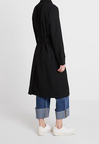 Person som bär en svart midjebelagd trenchcoat, uppvikta blå jeans med dragkedjor och vita sneakers, står mot en enfärgad vit bakgrund.