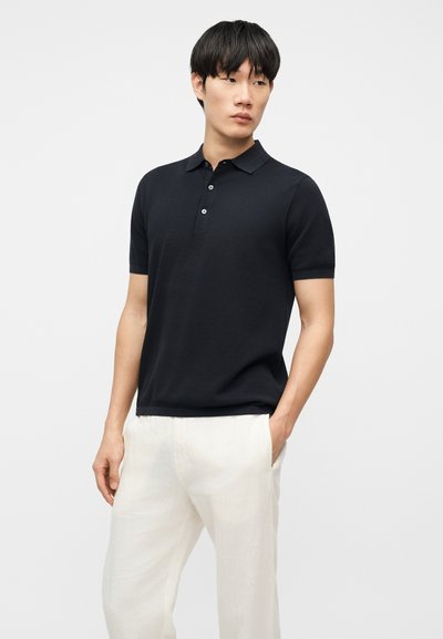 Homme debout, les mains dans les poches, portant un polo noir à manches courtes et un pantalon blanc sur un fond clair uni.