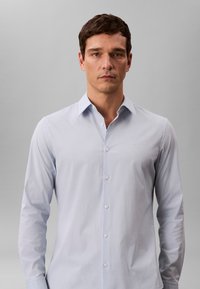 Helehall sinine pikkade varrukatega nööpidega särk, millel on terav krae, sile tekstuur ja rinnal peen logo, valgete nuppudega.