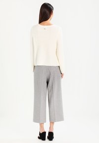 Kermanvärinen neulepaita, jossa on ribbineulos, yhdistetty korkeavyötäröisiin harmaisiin leveälahkeisiin housuihin. Asun täydentävät mustat korolliset kengät.