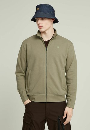 Chaqueta con cremallera de color verde oliva y bolsillos frontales, hecha de una tela suave. Combinada con un sombrero de cubo de mezclilla oscura que tiene un acento azul.