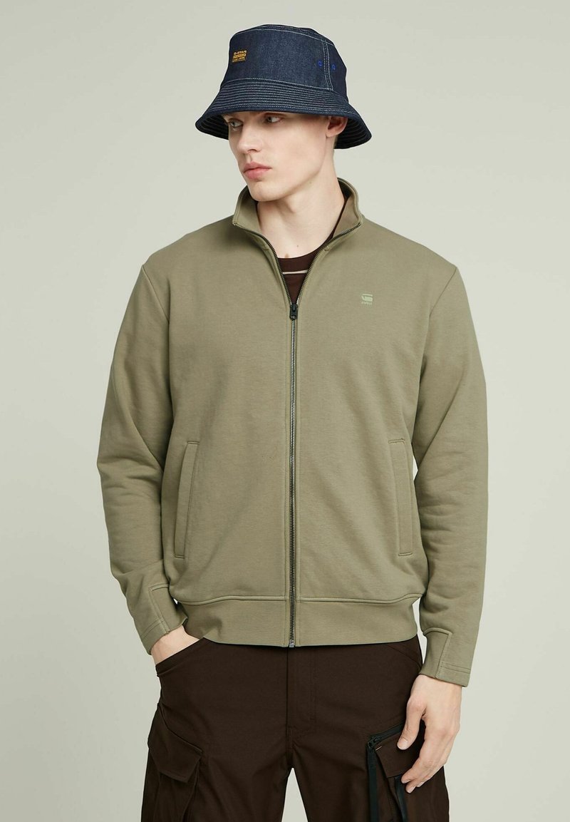 Giacca con zip verde oliva, dotata di tasche frontali e realizzata in tessuto morbido. Abbinata a un cappello a secchiello in denim scuro con un accento blu.