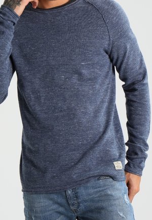 Chemise à manches longues bleu marine, à col rond, en tissu texturé, avec des manches raglan et une petite étiquette en bas à gauche.