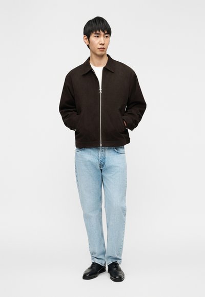 Νέος άνδρας που φοράει σκούρο καφέ μπουφάν με φερμουάρ, ανοιχτό μπλε τζιν και μαύρα παπούτσια, στέκεται με τα χέρια στις τσέπες μπροστά σε ένα απλό φόντο.