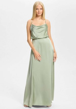 Blondynka stojąca w jasnooliwkowej satynowej, bezrękawowej sukni maxi na cienkich ramiączkach na jednolitym białym tle.