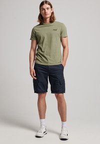 T-shirt verde oliva con scollatura rotonda, abbinato a pantaloni cargo blu scuro con tasche laterali, calze bianche e sneakers.