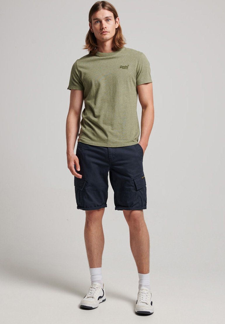 T-shirt verde oliva con scollatura rotonda, abbinato a pantaloni cargo blu scuro con tasche laterali, calze bianche e sneakers.