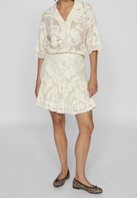 Camicia in pizzo crema con maniche corte, motivo decorativo; gonna in pizzo crema abbinata con orlo a volants; abbinata a ballerine maculate in nero e marrone.
